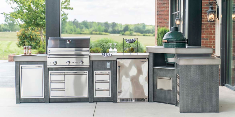 A linear outdoor kitchen with dark gray wood-textured cabinetry and a thick concrete countertop. It is equipped with a large stainless steel grill and a green "Big Green Egg" ceramic charcoal cooker integrated into the end of the unit.