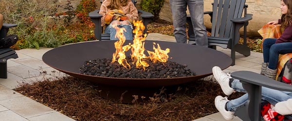 Family gathered around a large round fire pit on a stone patio with Adirondack chairs, blankets, and autumn plantings in the background.